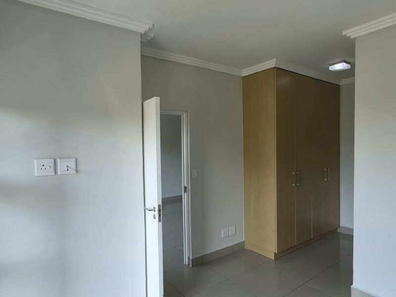 A bright, empty bedroom featuring neutral walls, tiled flooring, built-in wooden cupboards, and modern cornicing, offering a clean and versatile living space.