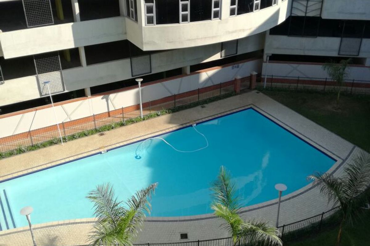 An elevated view of a communal swimming pool area featuring paved surrounds, landscaped garden, and perimeter fencing within an apartment complex.