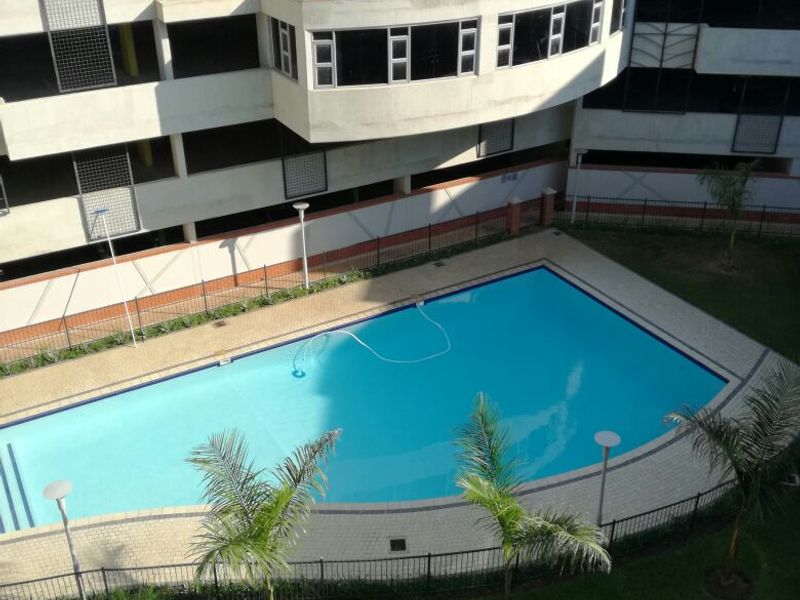 An elevated view of a communal swimming pool area featuring paved surrounds, landscaped garden, and perimeter fencing within an apartment complex.