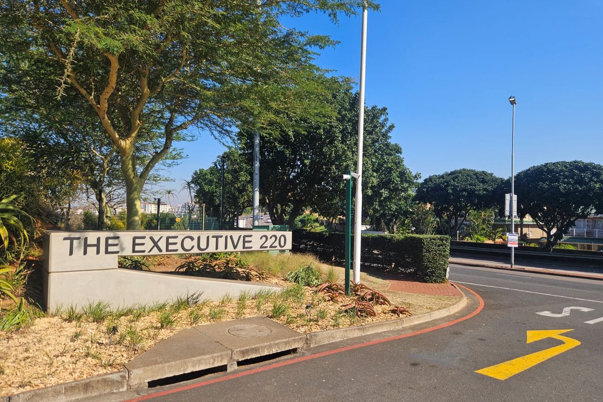 Street view of the entrance to The Executive 220, featuring a concrete signage wall, landscaped verge, and asphalt road access.