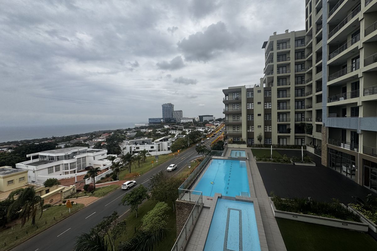 An elevated aerial view of the apartment complex featuring a swimming pool, landscaped grounds, and surrounding urban coastal landscape.
