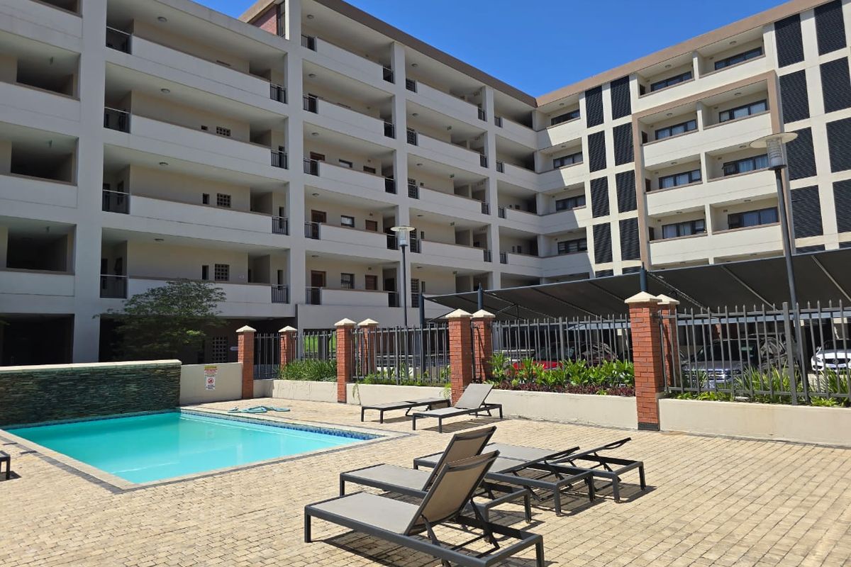 A well-maintained communal pool area featuring a paved deck, water feature, and secure perimeter fencing, set against a modern multi-story apartment building.
