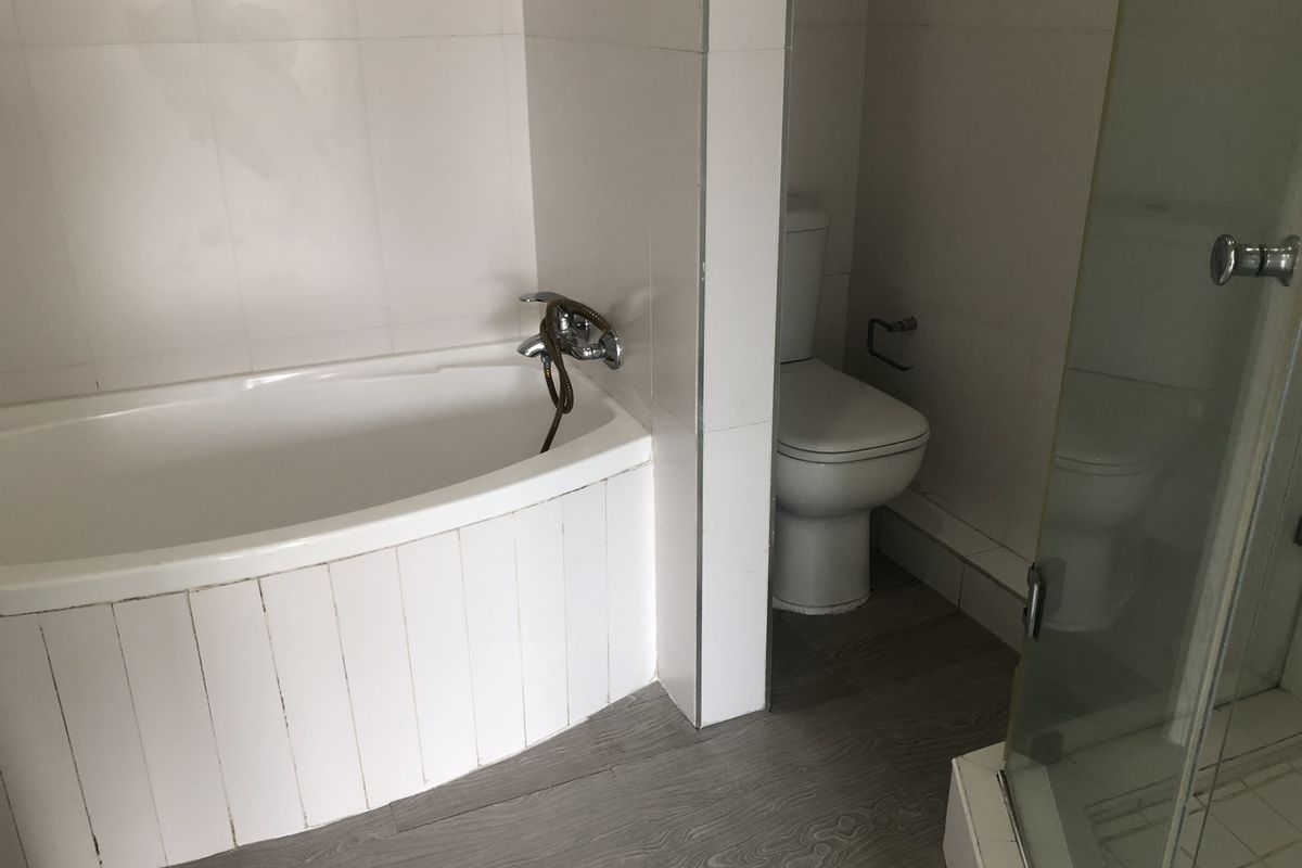 A functional bathroom featuring a bathtub, separate toilet area, and a glass-enclosed shower with neutral wall tiling and wood-look flooring.