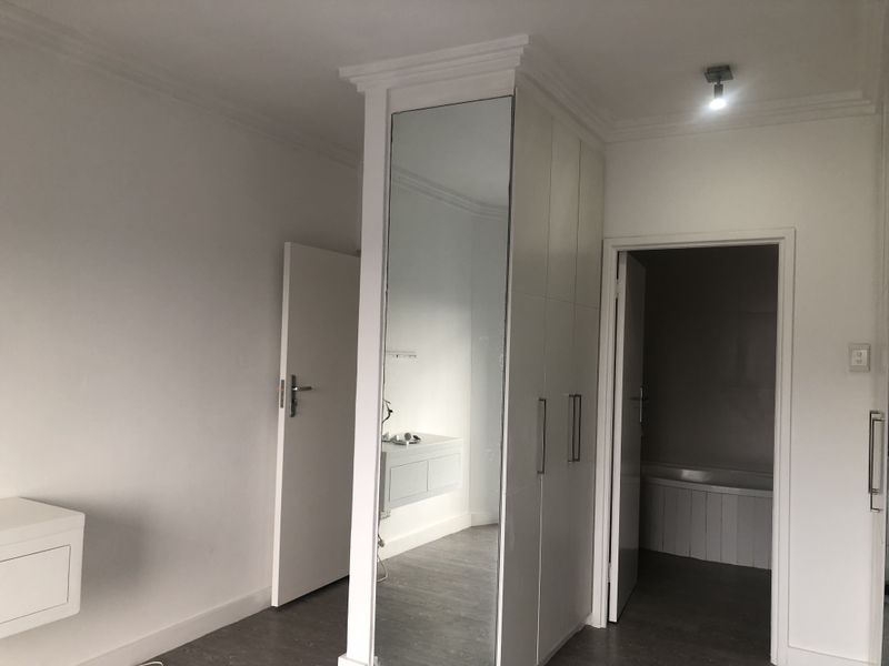 A bright, minimalist bedroom featuring built-in wardrobes with a mirrored panel, crown molding, and direct access to an adjoining bathroom.