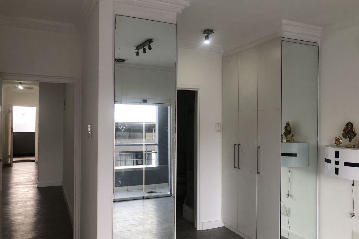 A clean bedroom featuring built-in cupboards with mirrored doors, laminate flooring, and modern ceiling lighting, leading to an adjacent balcony area.