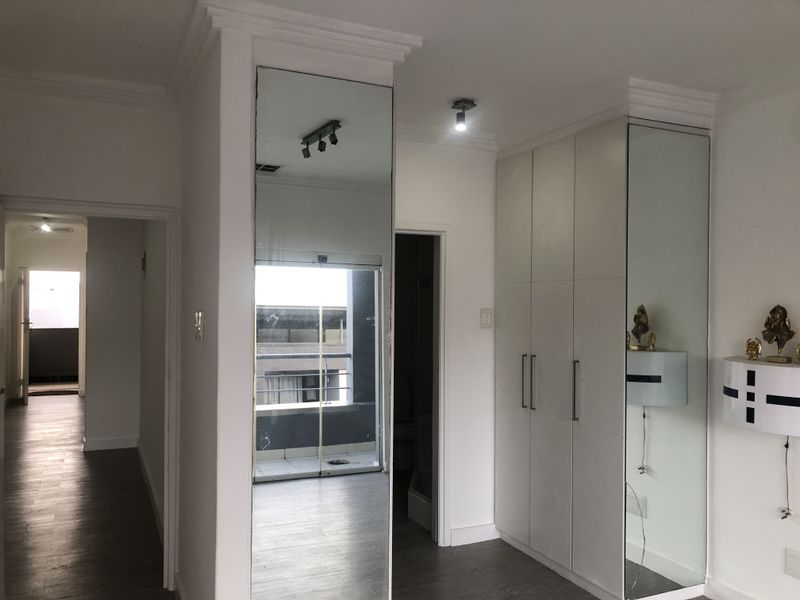 A clean bedroom featuring built-in cupboards with mirrored doors, laminate flooring, and modern ceiling lighting, leading to an adjacent balcony area.