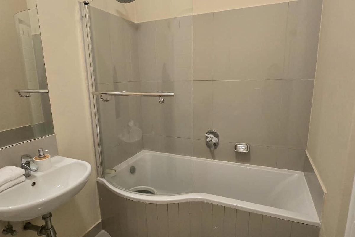 A clean, functional bathroom featuring a wall-mounted basin, a bathtub with a glass shower screen, and neutral wall tiling.