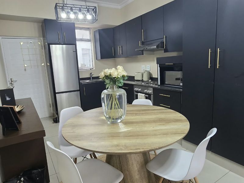 Modern open-plan kitchen and dining area featuring sleek dark cabinetry, integrated appliances, and contemporary pendant lighting with neutral tiled flooring.