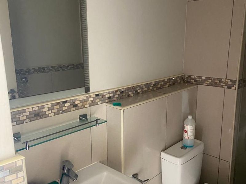 A functional bathroom featuring a pedestal washbasin, wall-mounted mirror, glass shelving, and neutral ceramic tiling with a decorative mosaic border.