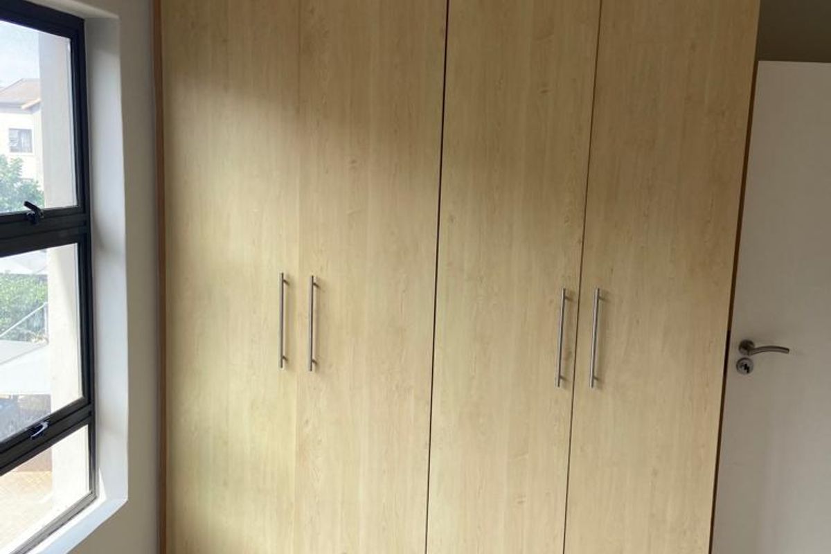 A bright bedroom featuring light wood-finish built-in wardrobes, neutral tiled flooring, and a modern aluminium-framed window.