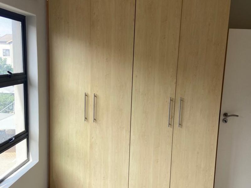 A bright bedroom featuring light wood-finish built-in wardrobes, neutral tiled flooring, and a modern aluminium-framed window.