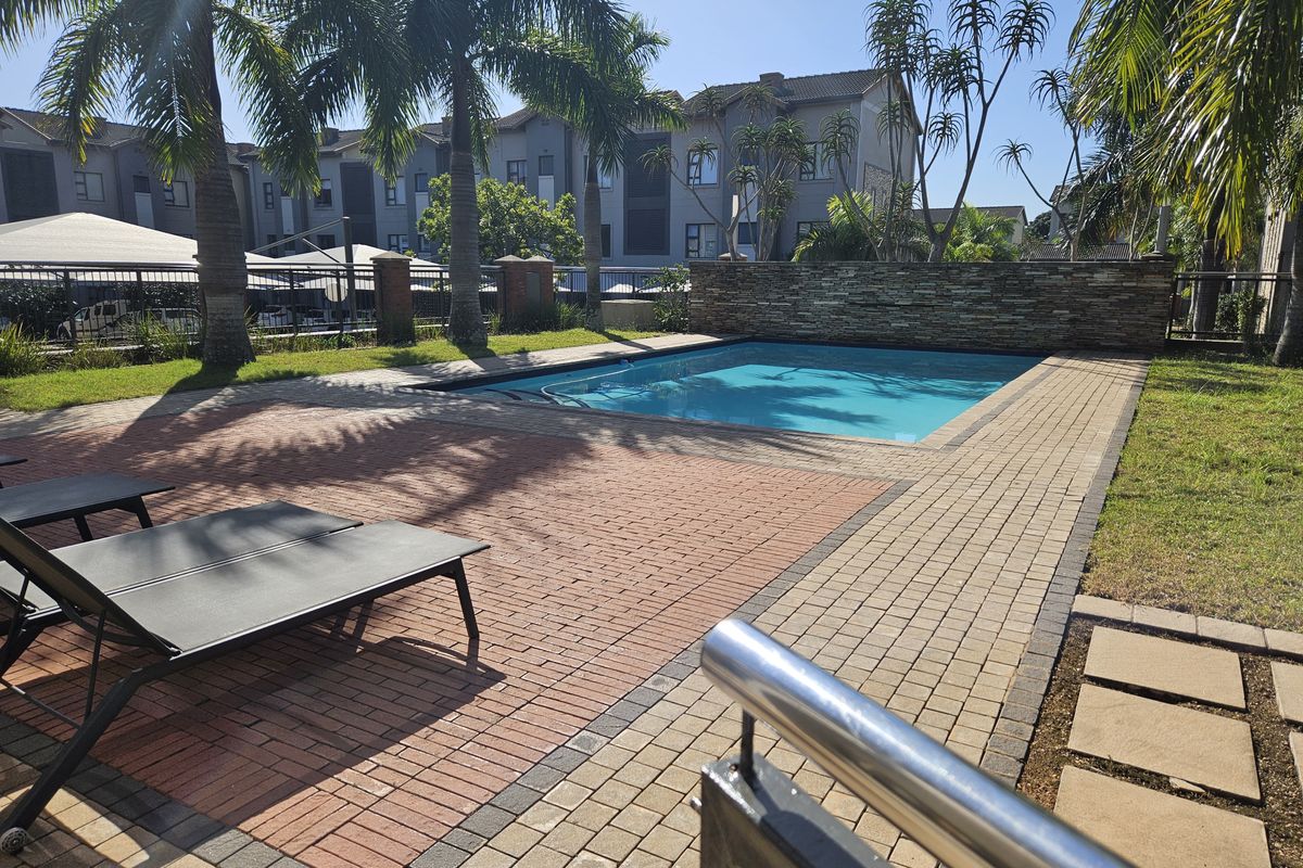 A well-maintained communal swimming pool area featuring a stone feature wall, paved decking, and surrounding landscaped gardens within an apartment complex.
