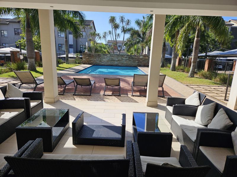 A well-maintained outdoor pool area featuring a swimming pool, stone feature wall, and a spacious paved patio under a covered structure.