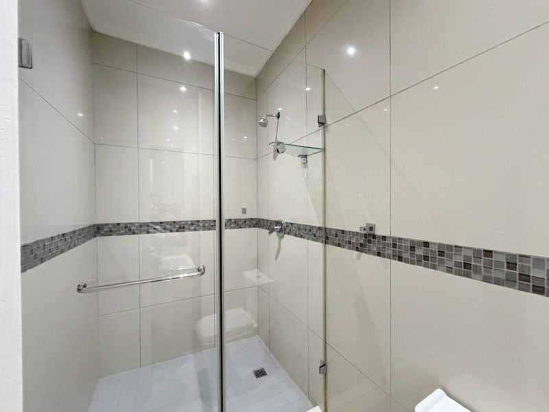 A clean, modern bathroom shower featuring neutral wall tiles with a decorative mosaic border, glass enclosure, and recessed lighting.