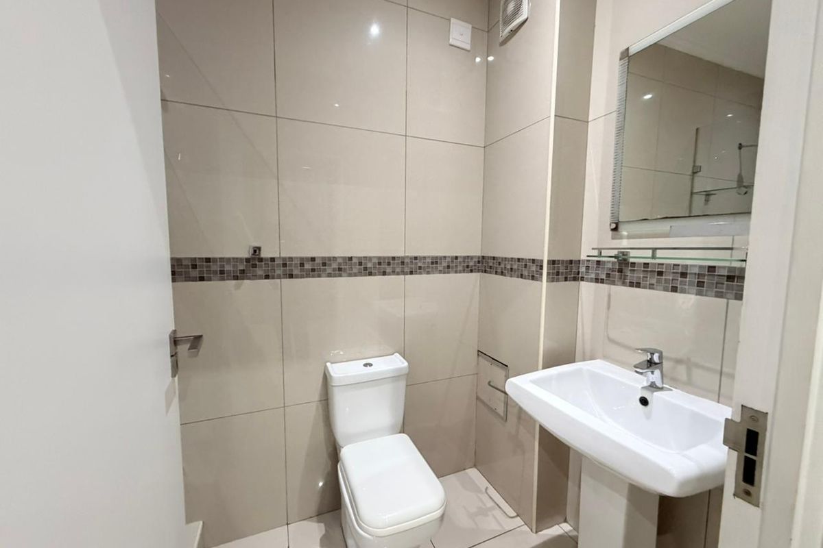 A clean and modern bathroom featuring a pedestal washbasin, toilet, and neutral wall tiling with a decorative mosaic accent border.