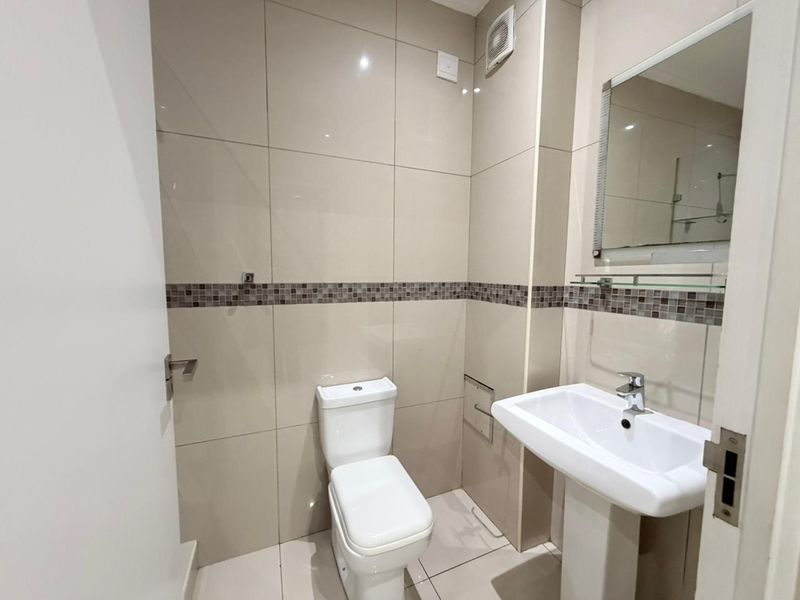 A clean and modern bathroom featuring a pedestal washbasin, toilet, and neutral wall tiling with a decorative mosaic accent border.