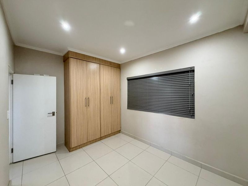 A clean, empty bedroom featuring light wood-finish built-in cupboards, neutral tiled flooring, and modern recessed downlighting.