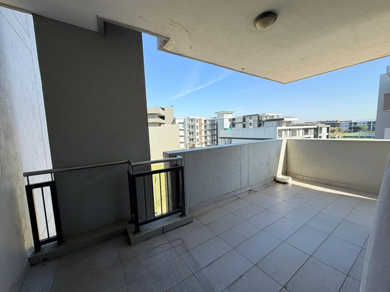 A spacious covered balcony featuring tiled flooring and a metal balustrade, offering an open view of the surrounding apartment complex.