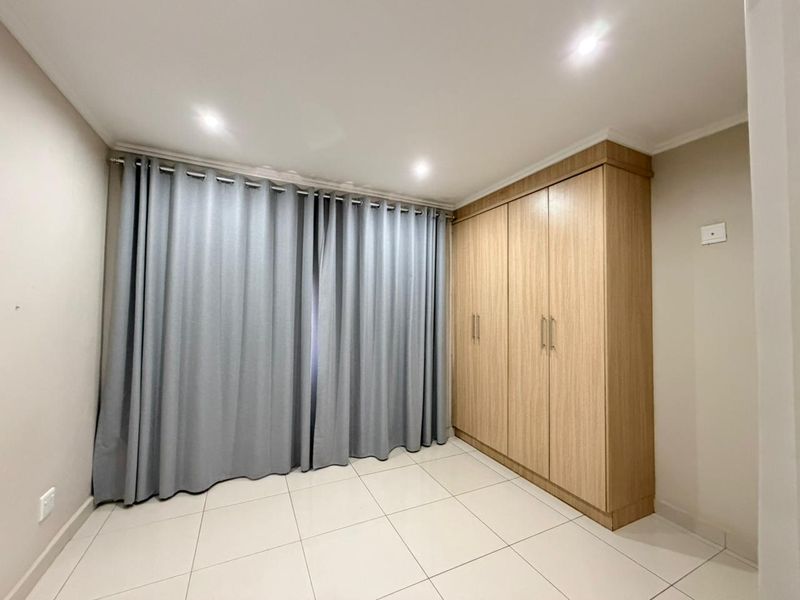 A bright, empty bedroom featuring neutral tiled flooring, recessed downlighting, and a large built-in wooden wardrobe with ample storage space.