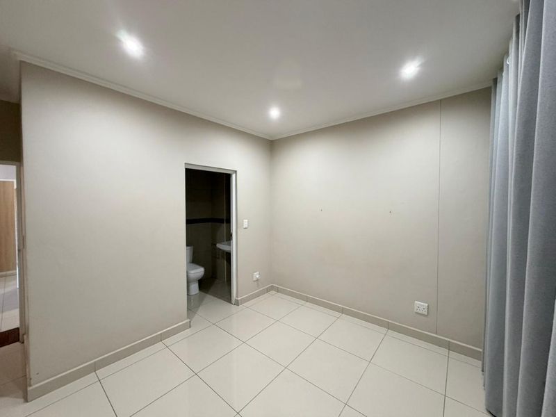 A clean, empty bedroom featuring neutral tiled flooring, recessed downlighting, and direct access to an en-suite bathroom.