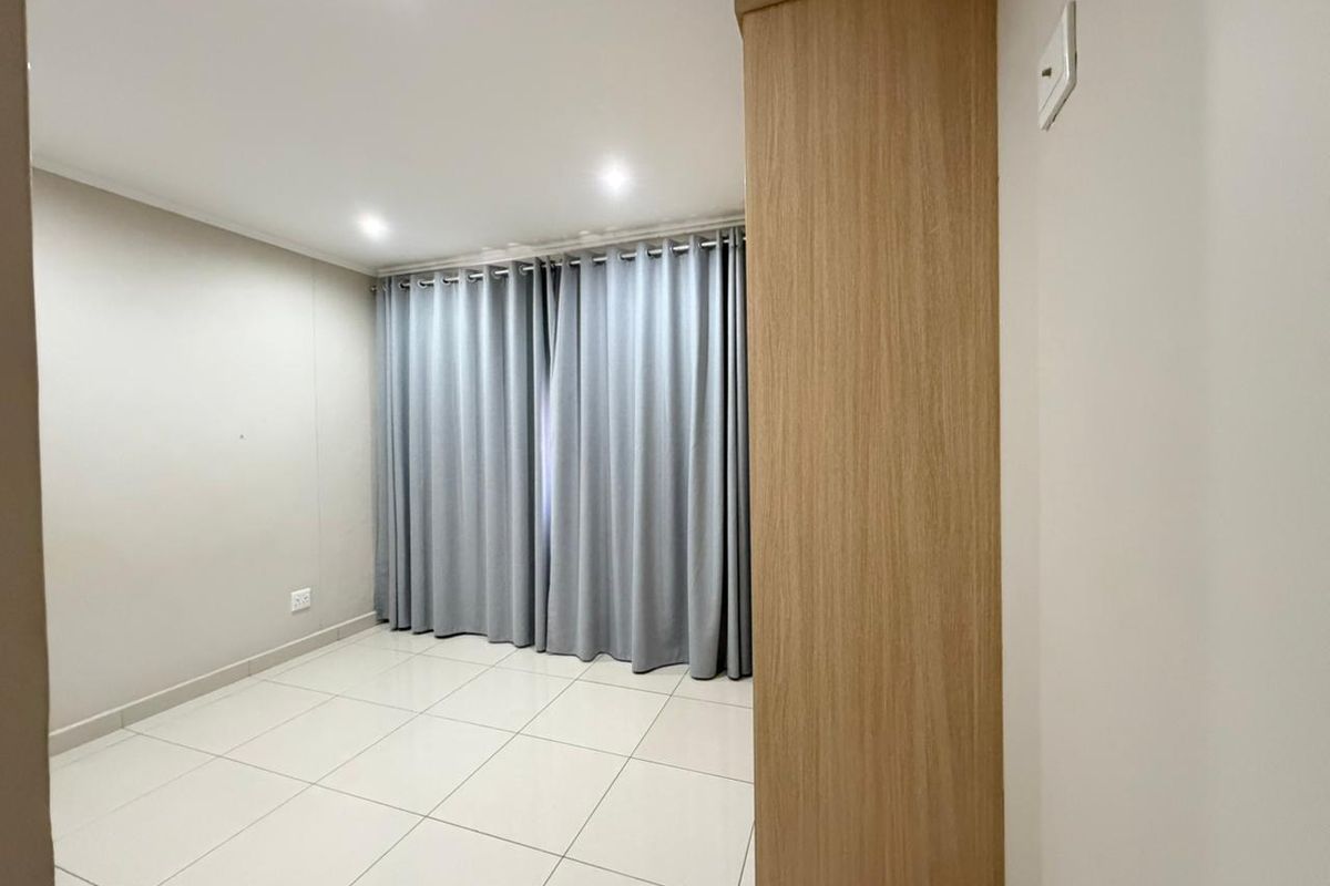 A bright, empty bedroom featuring neutral tiled flooring, recessed downlighting, and a built-in wooden cupboard, offering a clean and versatile living space.
