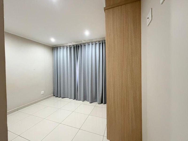 A bright, empty bedroom featuring neutral tiled flooring, recessed downlighting, and a built-in wooden cupboard, offering a clean and versatile living space.