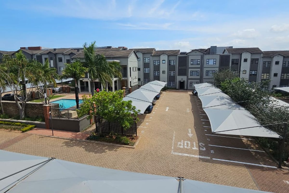 Aerial view of the apartment complex featuring a communal swimming pool, paved parking area with shade-cloth carports, and landscaped grounds.