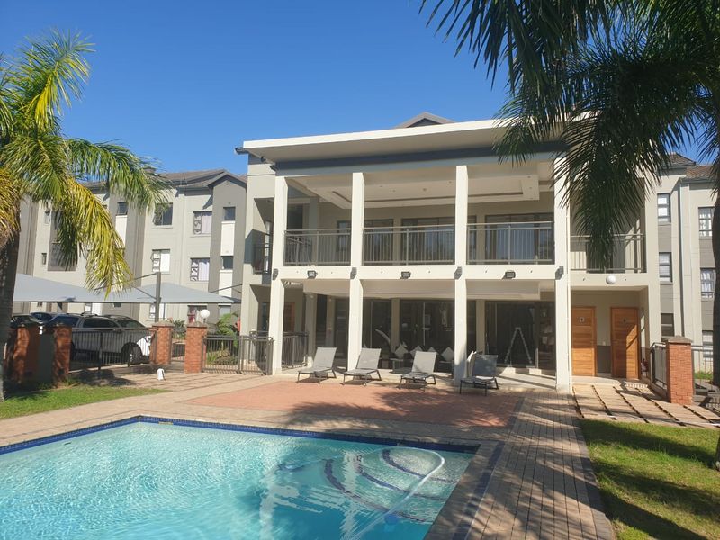 A well-maintained apartment complex pool area featuring a paved deck, covered patio, and balcony overlooking the swimming pool.