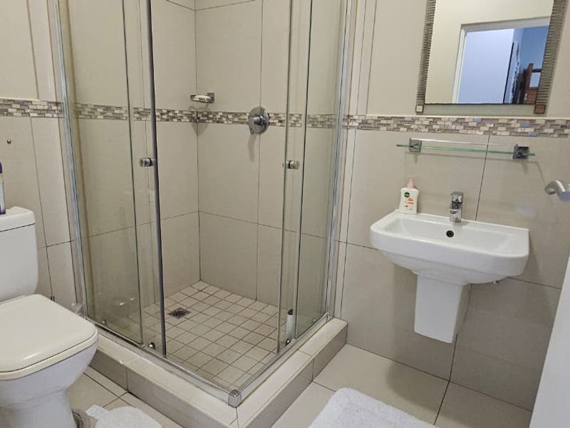 A clean, functional bathroom featuring a glass-enclosed shower, pedestal basin, and toilet with neutral wall and floor tiling.