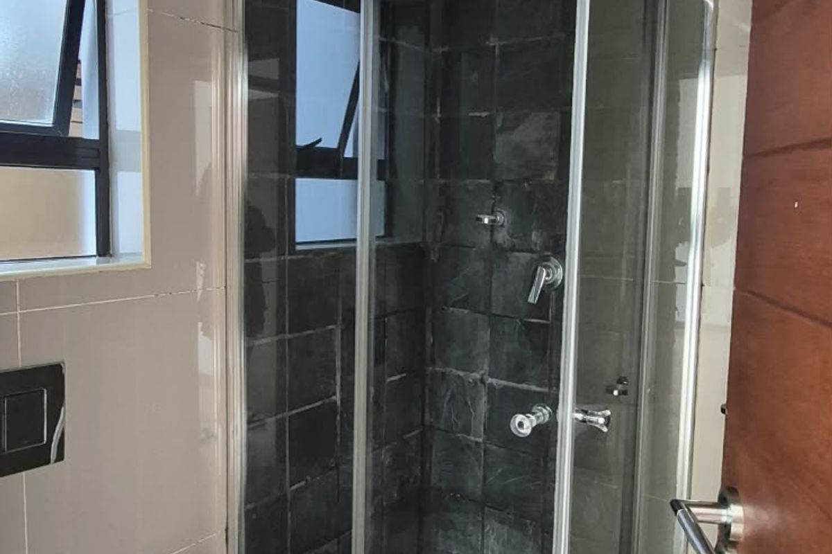 A compact bathroom featuring a corner shower enclosure with dark tiling, a wall-mounted toilet, and neutral floor tiles.