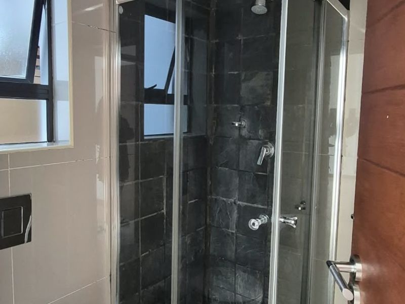 A compact bathroom featuring a corner shower enclosure with dark tiling, a wall-mounted toilet, and neutral floor tiles.