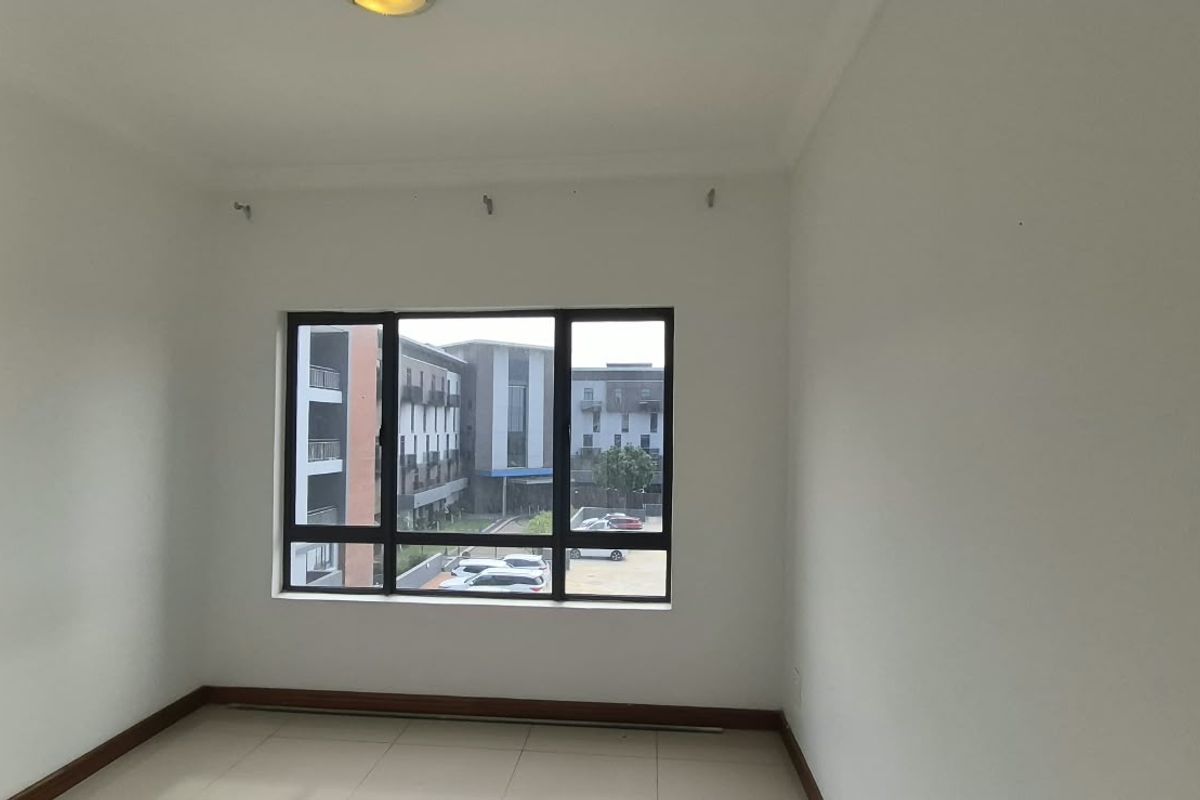 A bright, empty bedroom featuring tiled flooring and a large aluminium-framed window with views of the surrounding apartment complex.