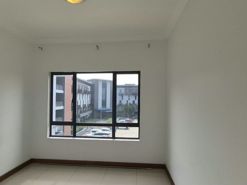 A bright, empty bedroom featuring tiled flooring and a large aluminium-framed window with views of the surrounding apartment complex.