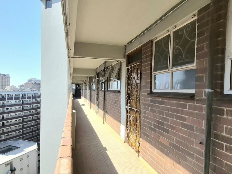 An exterior balcony walkway featuring brick walls, security gates, and tiled flooring, overlooking the surrounding urban apartment complex.