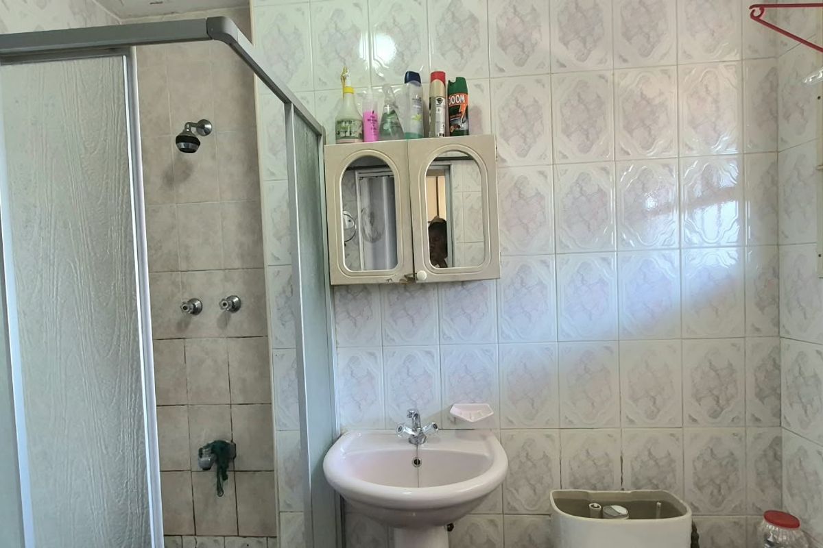 A functional bathroom featuring a shower enclosure, pedestal basin, and toilet, finished with wall tiles and a window with security bars.