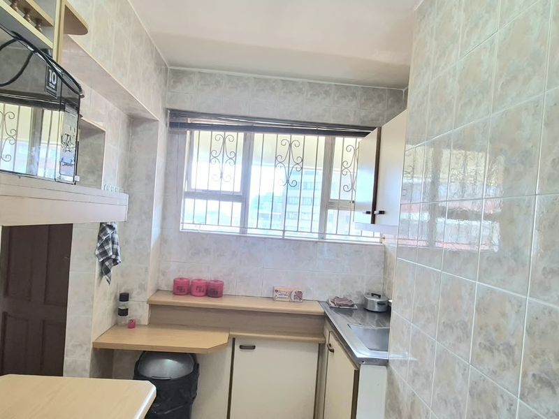 A compact kitchen featuring dated wall tiles, built-in cabinetry, a stainless steel sink, and an extractor fan, with natural light from a window.