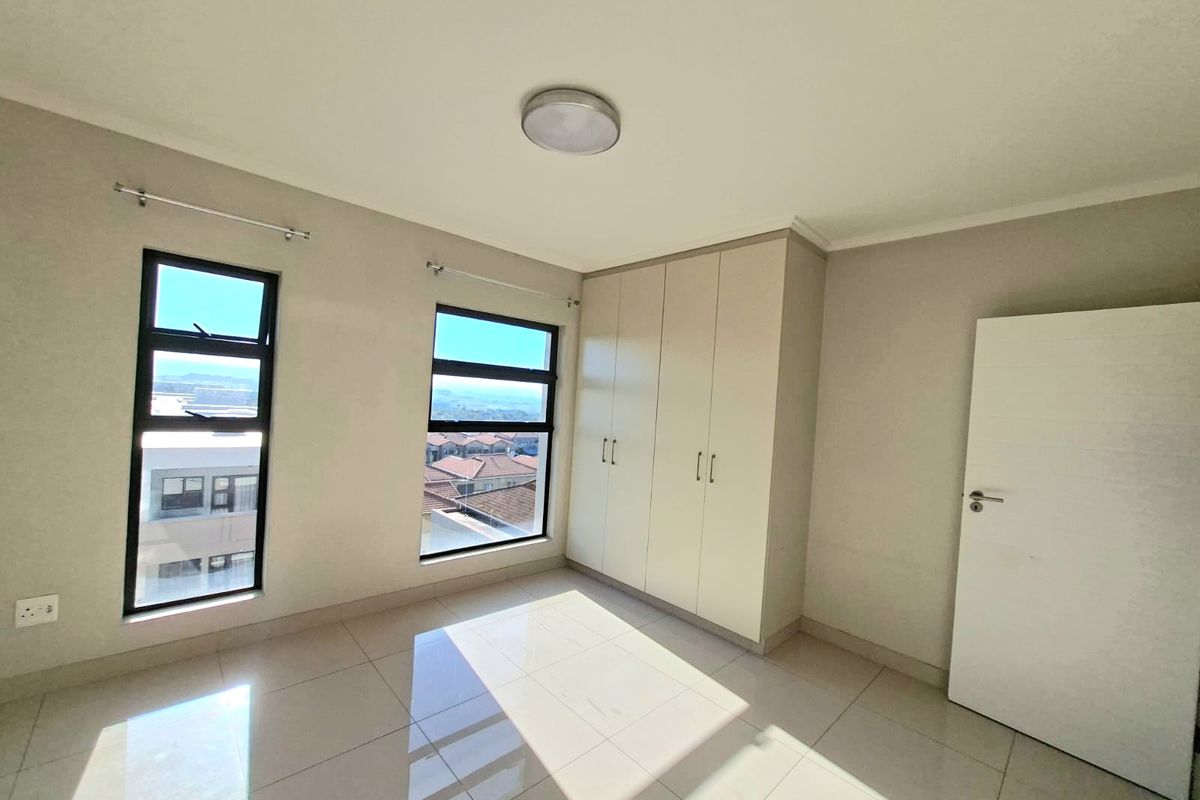 A bright, empty bedroom featuring neutral tiled flooring, built-in cupboards, and large aluminium-framed windows offering natural light and neighbourhood views.