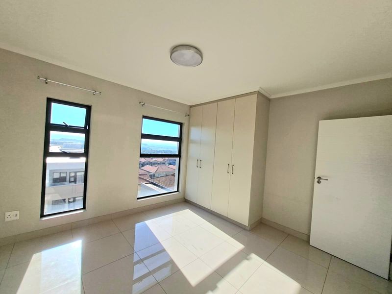A bright, empty bedroom featuring neutral tiled flooring, built-in cupboards, and large aluminium-framed windows offering natural light and neighbourhood views.