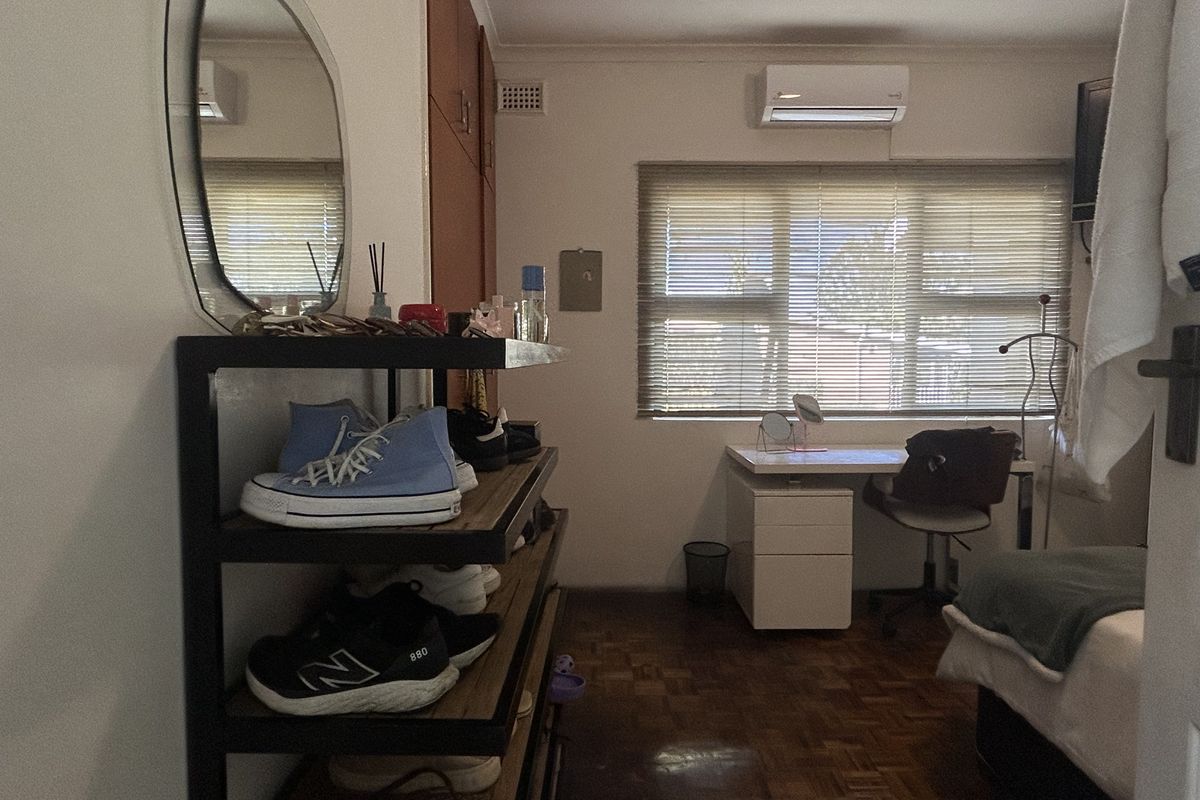 A bedroom featuring parquet flooring, built-in cupboards, and an air conditioning unit, with natural light filtered through venetian blinds.