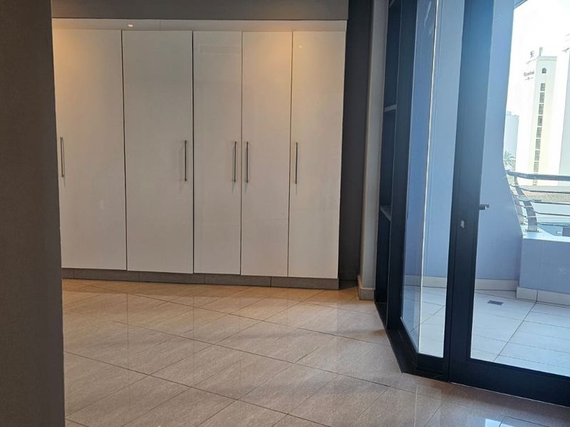 A bright, empty bedroom featuring modern white built-in cupboards, tiled flooring, and direct access to a private balcony through a large sliding glass door.