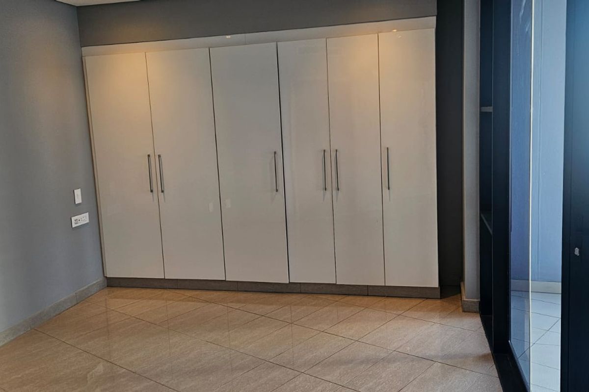A bright, empty bedroom featuring modern built-in cupboards, tiled flooring, and a sliding glass door leading to an outdoor balcony area.