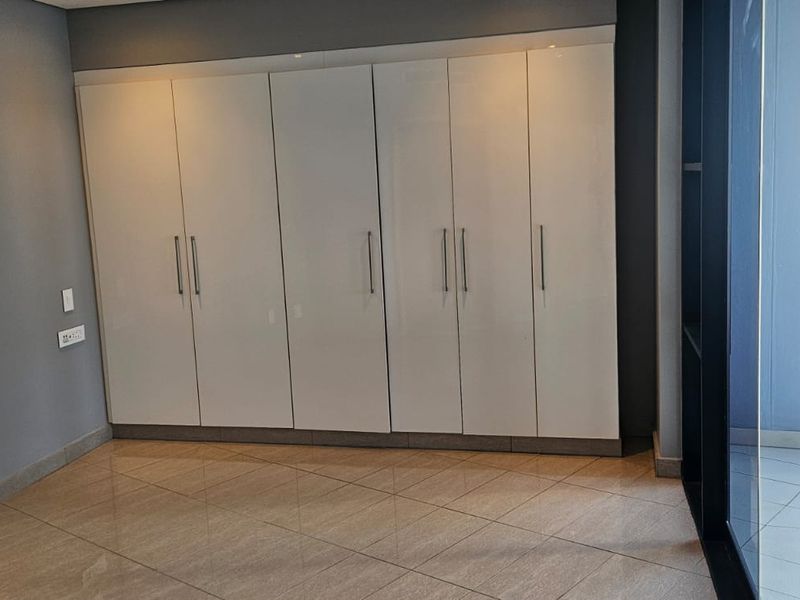A bright, empty bedroom featuring modern built-in cupboards, tiled flooring, and a sliding glass door leading to an outdoor balcony area.