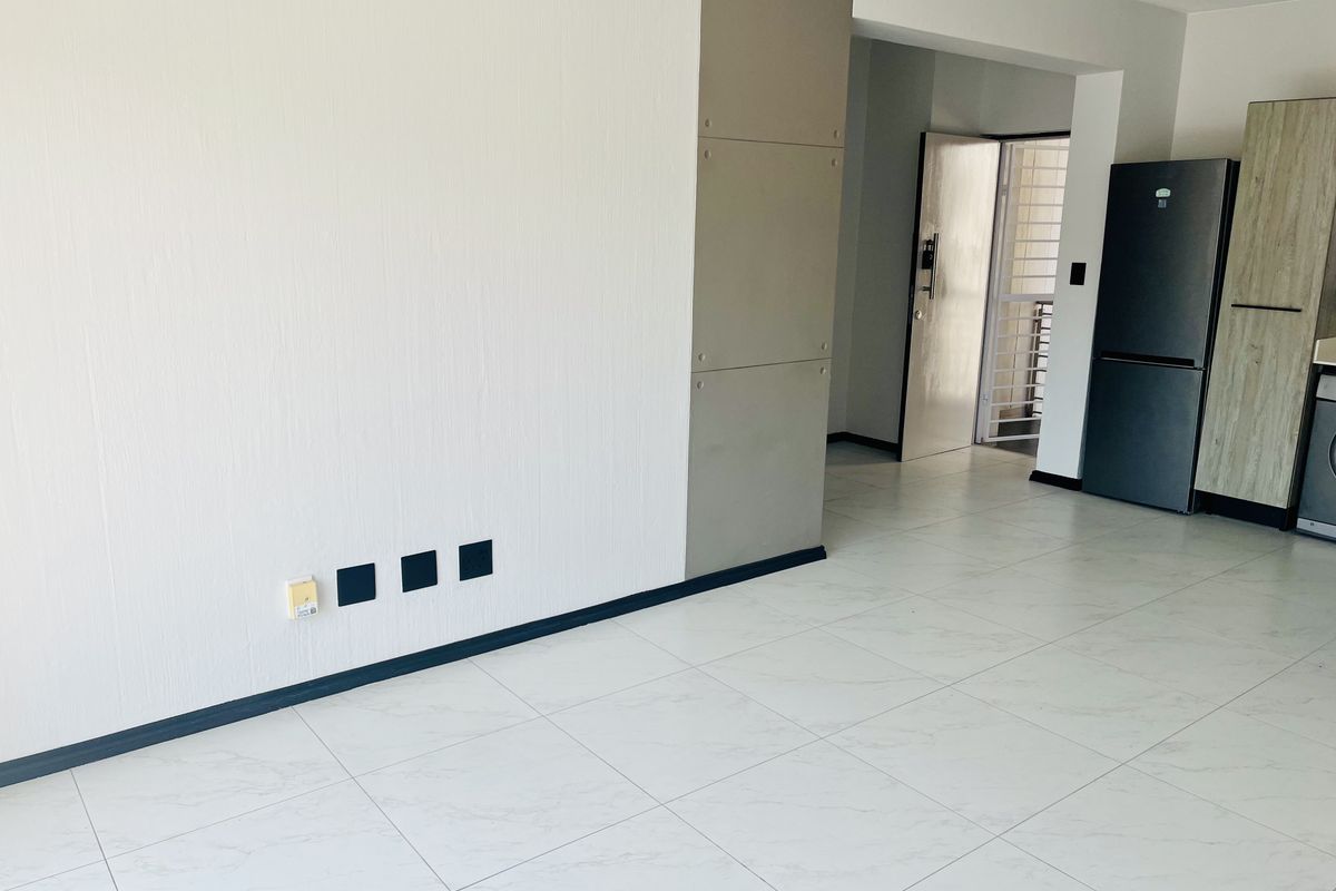 An open-plan living area featuring modern white tiled flooring, neutral walls, and built-in kitchen cabinetry, leading to a secure entrance.