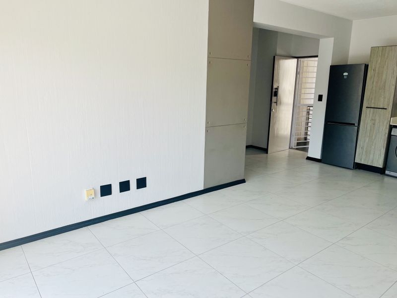 An open-plan living area featuring modern white tiled flooring, neutral walls, and built-in kitchen cabinetry, leading to a secure entrance.