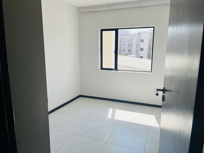 A bright, empty bedroom featuring modern white tiled flooring, neutral walls, and a black-framed window providing natural light.