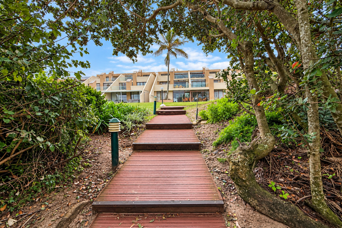 A timber boardwalk path leads through a landscaped garden towards the rear elevation of the apartment complex.