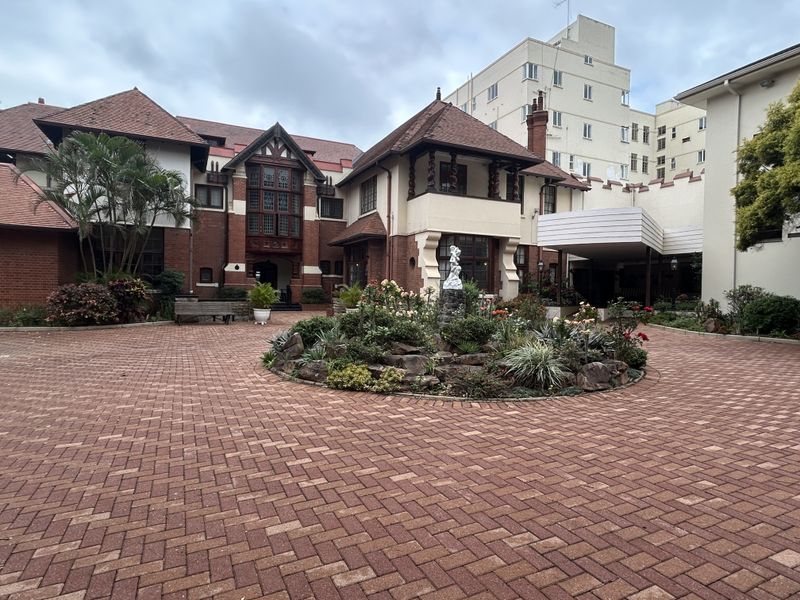 A well-maintained retirement village entrance featuring a brick-paved courtyard, central landscaped garden with a stone fountain, and traditional architectural detailing.