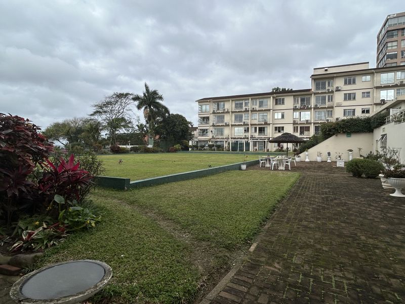 A view of the rear exterior of the retirement village, featuring a paved courtyard area, a large landscaped lawn, and the multi-story residential building.