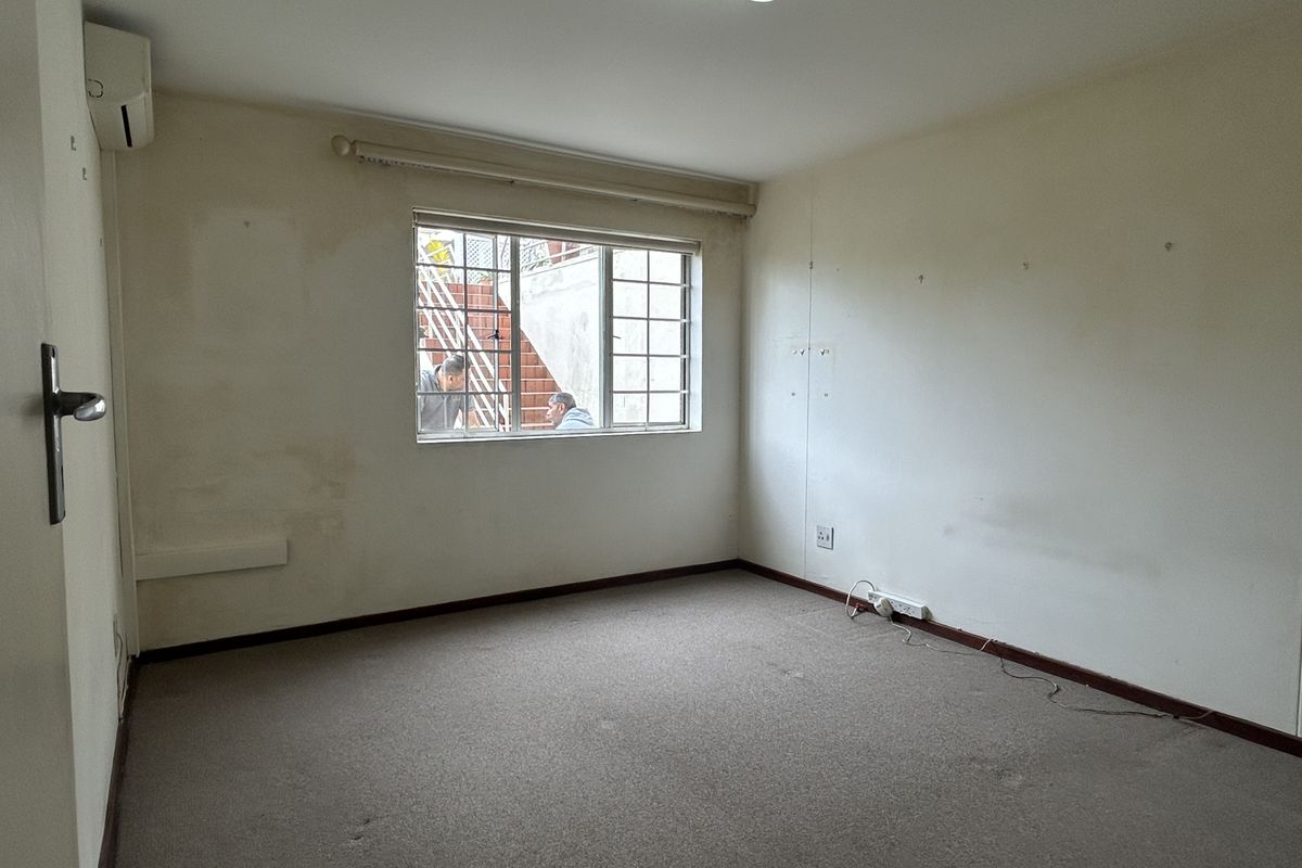 An empty bedroom featuring carpeted flooring, a wall-mounted air conditioning unit, and a window with security bars, requiring cosmetic updates.