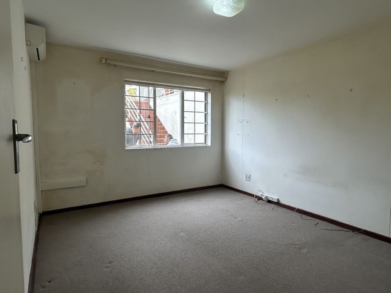 An empty bedroom featuring carpeted flooring, a wall-mounted air conditioning unit, and a window with security bars, requiring cosmetic updates.